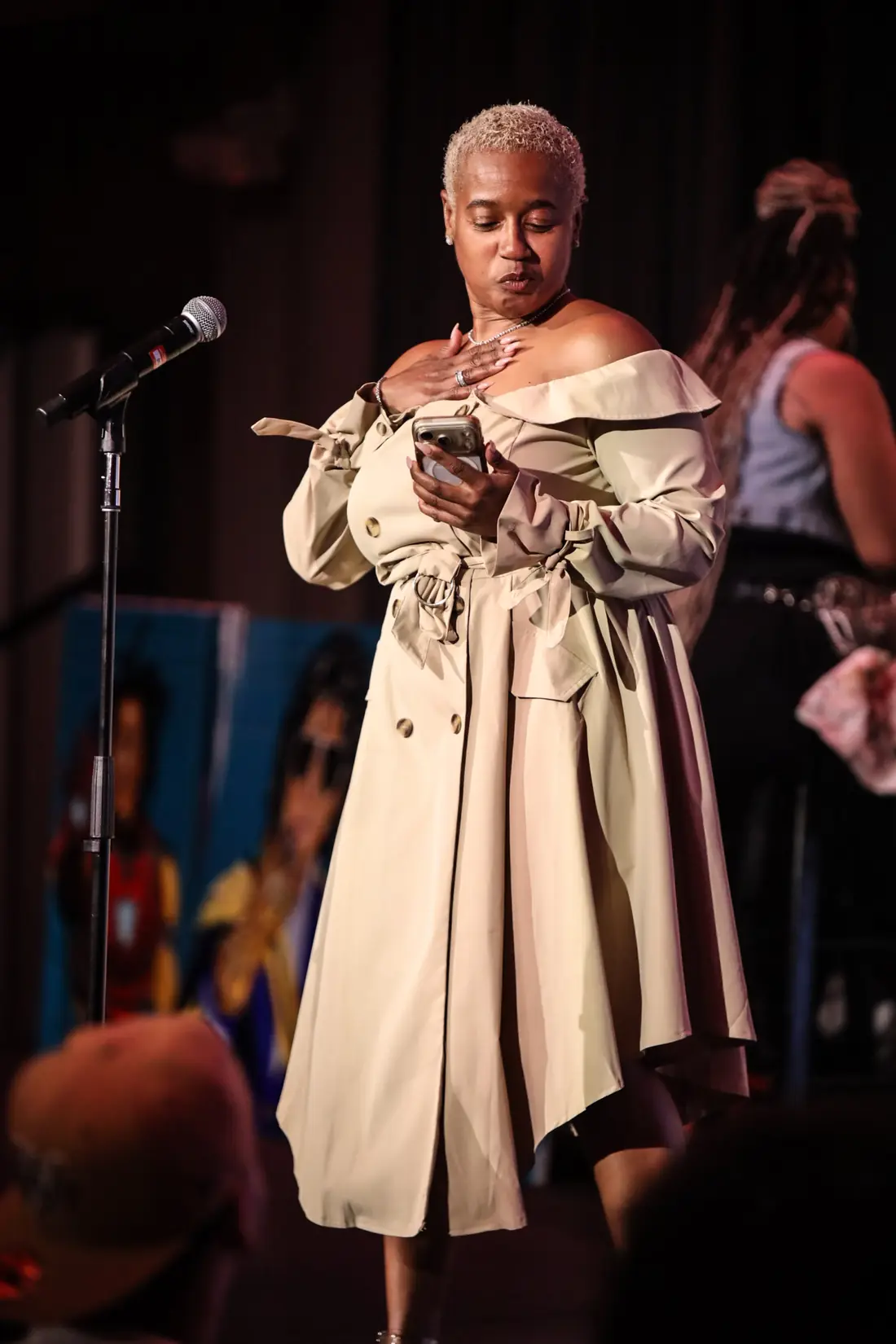 A poet in a trench coat reading from her phone at the mic